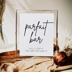 Puede incluir: Un letrero blanco en un marco dorado con las palabras "parfait bar" en escritura negra. El letrero también incluye el texto "¡Rellénalo, cúbrelo, como quieras!" Elementos decorativos: hierba de la pampa y una bandeja de madera.
