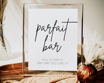 May include: A white sign in a gold frame with the words "parfait bar" in black script. The sign also includes the text "Fill it, top it, any way you like it!" Decorative elements include pampas grass and a wooden tray.