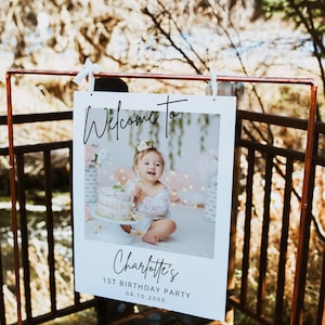 May include: White welcome sign with a photo of a baby girl in a white dress. The sign says "Welcome to Charlotte's 1st Birthday Party 04.10.20XX".
