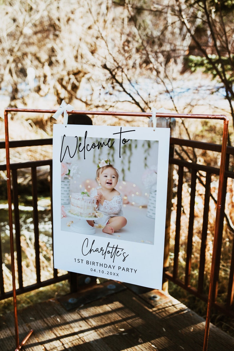 May include: White welcome sign with a photo of a baby girl in a white dress. The sign says "Welcome to Charlotte's 1st Birthday Party 04.10.20XX".