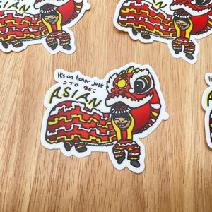 Könnte beinhalten: Ein Aufkleber mit einer Cartoon-Illustration einer Person in einem roten und gelben Löwentanzkostüm. Der Text auf dem Aufkleber lautet "Es ist eine Ehre, einfach ASIAN zu sein".