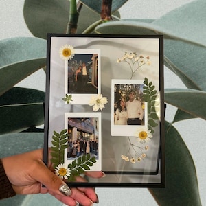 May include: A rectangular black metal floating frame containing three square white-bordered photographs. The display is decorated with pressed white daisy flowers, small white baby's breath sprigs, and green fern leaves arranged between the photos.