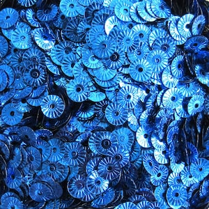 May include: A close-up view of a pile of shiny, blue sequins. Each sequin is round with a central hole and a radial pattern, resembling small, metallic flowers. The sequins are scattered, creating a textured, reflective surface.