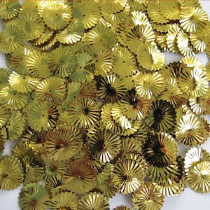 May include: A pile of small, shiny, gold-coloured paper fan embellishments. Each fan has a circular centre with radiating, pleated edges. The fans are scattered, creating a textured, decorative effect, suitable for crafts and scrapbooking.