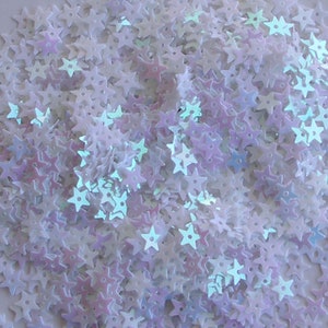May include: A close-up of a pile of iridescent star-shaped sequins. The sequins are mainly white with touches of pink, purple, and blue, creating a shimmering effect. These decorative embellishments are ideal for crafts and adding sparkle.