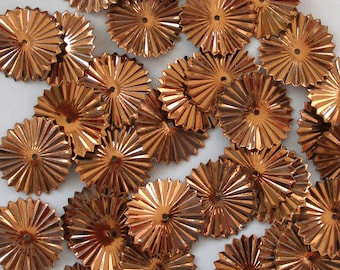 May include: A collection of small, copper-colored metal embellishments. Each piece has a circular shape with a central hole and radiating, ridged edges, resembling miniature sunbursts. The pieces are scattered across a white surface.