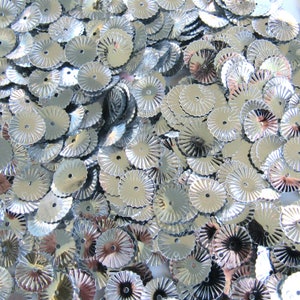 May include: A pile of shiny, silver sequins. Each sequin has a scalloped edge and a small hole in the center. The sequins are arranged closely together, creating a reflective surface.