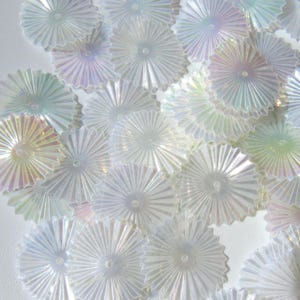 May include: A collection of iridescent, fan-shaped embellishments. These decorative pieces have a radial design with a central point and are in shades of white, with hints of pastel colors like pink, blue, and green. They appear to be made of a translucent material.