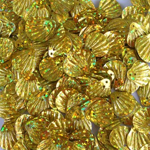May include: A close-up of a collection of gold-coloured sequins. The sequins are shaped like stylised shells with a textured, fan-like design. Each sequin has a small hole for sewing. The sequins have a shiny, iridescent finish.