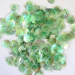 May include: A collection of iridescent green seashell-shaped sequins. The sequins have a fan-like design with a hole at the top for stringing. The light reflects off the sequins, creating a shimmering effect. These craft supplies are ideal for embellishing clothing or accessories.