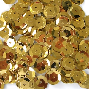 May include: A pile of shiny, gold-colored sequins. Each sequin is a flat, hexagonal shape with a small hole in the center. The sequins are scattered across a white surface, reflecting light and creating a sparkling effect. Ideal for crafts and embellishments.