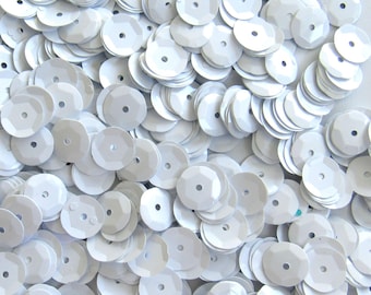May include: A close-up view of a pile of white, faceted sequins. Each sequin is round with a central hole and a multi-faceted surface, creating a shimmering effect. The sequins are scattered, overlapping each other, and filling the frame.