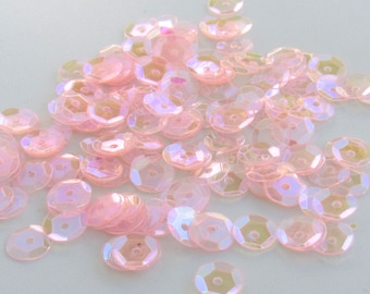Puede incluir: Un montón de lentejuelas rosas iridiscentes. Cada lentejuela es redonda con un agujero hexagonal en el centro. Las lentejuelas tienen una superficie brillante y reflectante, creando un efecto brillante. El color cambia de rosa a amarillo pálido.