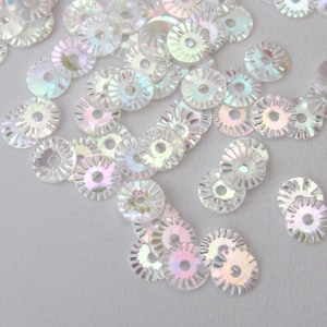 Puede incluir: Un conjunto de lentejuelas iridiscentes y transparentes. Cada lentejuela es circular con un agujero central y un borde festoneado. Las lentejuelas reflejan un arcoíris de colores, incluyendo rosa, azul y verde, creando un efecto brillante. Ideal para proyectos de manualidades.