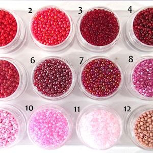 May include: Twelve clear plastic jars filled with small round beads in various shades of red, pink, and beige. Each jar is labeled with a black number from 1 to 12, arranged in three rows of four, showcasing a gradient of colors from bright red to light pink.