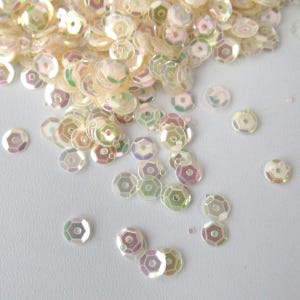 May include: A collection of iridescent sequins in different sizes and shapes. The sequins are mainly a pale, creamy colour with touches of pink, green, and purple, creating a shimmering effect. Each sequin has a central hole.