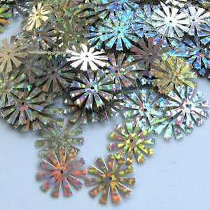 May include: A collection of silver, star-shaped sequins with a holographic, iridescent finish. Each sequin has a central hole and radiating points, creating a sparkling, decorative element. The sequins are scattered, suggesting their use in crafts or embellishments.