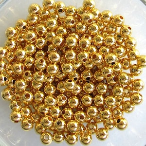 May include: A collection of small, shiny, gold-colored beads. Each bead is spherical with a small hole running through the center. The beads are densely packed together, reflecting light and creating a bright, textured surface. Ideal for jewelry making and crafts.