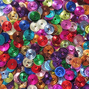 Puede incluir: Primer plano de un montón de lentejuelas de colores. Las lentejuelas son redondas con un agujero en el centro y son de varios colores, incluyendo rojo, azul, verde, morado, dorado y plateado. Las lentejuelas están dispersas y superpuestas.