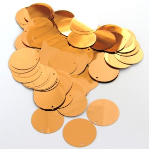May include: A collection of shiny, metallic copper-colored sequins scattered across a white surface. The sequins are round with a small hole near the edge, ideal for sewing or crafting projects. The sequins vary in size and are reflective.