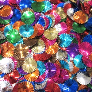 May include: A close-up view of a collection of colorful, metallic, fan-shaped embellishments. The decorations are in various colors, including blue, green, orange, pink, purple, and gold. Each piece has a small hole in the center.