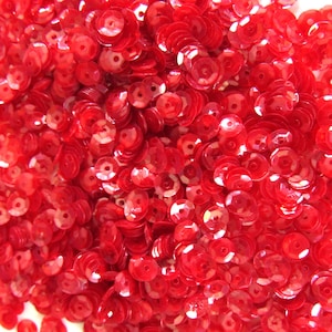 May include: A close-up image displays a mass of red sequins. These small, disc-shaped embellishments are scattered, creating a textured surface. Each sequin has a central hole, and they reflect light, adding a sparkling effect. Suitable for craft projects.