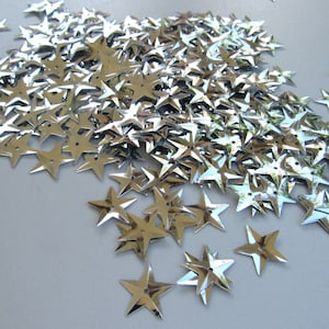 May include: A collection of shiny, silver star-shaped sequins scattered across a light gray surface. Each sequin has multiple points and a small hole in the center. The sequins reflect light, creating a sparkling effect.