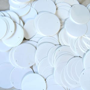 May include: A collection of white, circular paper tags with small holes near the top. The tags are stacked and scattered, creating a textured, layered effect. The tags are blank, offering space for customization.
