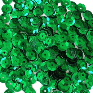 May include: A close-up shot of a pile of shiny, emerald green sequins. Each sequin is a flat, hexagonal disc with a small hole in the center. The sequins are scattered, creating a textured, reflective surface.