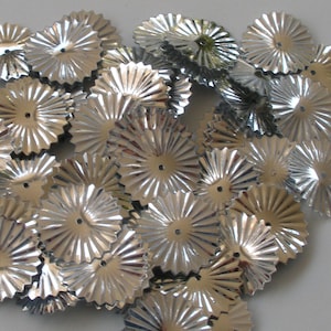 May include: A pile of silver, starburst-shaped paper decorations. Each piece has a central hole and radiating lines, creating a textured, metallic appearance. The decorations are scattered, suggesting a craft or party supply.