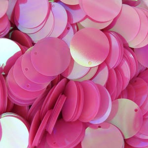 Op de afbeelding: Close-up van een stapel roze, iriserende pailletten. De pailletten zijn rond met een klein gaatje aan de rand. Ze hebben een glanzend, reflecterend oppervlak en zijn willekeurig verspreid.