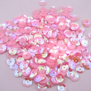 May include: A pile of pink sequins with a shimmering, iridescent finish. The sequins are round with a hole in the center, and they vary slightly in color, ranging from light to darker shades of pink. These decorative embellishments are ideal for crafts and sewing projects.