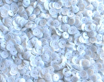 May include: A close-up shot of a pile of white sequins. The sequins are disc-shaped with a hole in the center, and they are scattered across the frame. The sequins have a slightly iridescent sheen, reflecting the light.