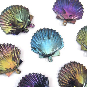 Pode incluir: Vários pingentes de concha iridescentes em tons de azul, roxo e dourado. Cada pingente tem uma borda ondulada e um pequeno laço para prender a joias ou outros trabalhos manuais. Dispostos sobre um fundo branco.