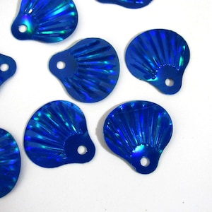 Könnte beinhalten: Nahaufnahme von mehreren irisierenden blauen muschelförmigen Pailletten. Jede Paillette hat ein fächerartiges Design mit einem kleinen Loch oben zum Annähen. Die Pailletten sind auf einem weißen Hintergrund verstreut.