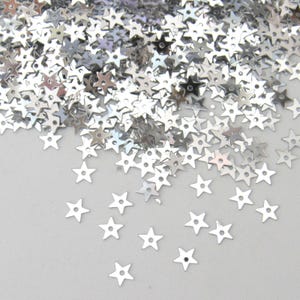 May include: A collection of small, metallic silver star-shaped sequins. Each individual star features a flat, reflective surface with a single, centred circular hole, designed for attachment or embellishment in various craft and decorative projects.