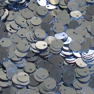 以下が含まれることがあります： キラキラ光る銀色のスパンコールの山をクローズアップ。各スパンコールは丸く、上部に小さな穴があります。スパンコールは散らばっており、テクスチャのある表面を作り出しています。手芸や装飾に最適です。