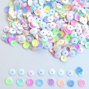 May include: A collection of iridescent, round sequins in various pastel colors, including pink, yellow, blue, and green. Each sequin has a small hole in the center. The sequins are scattered across a white surface, with some arranged in a neat row.