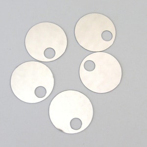 May include: Five circular silver-colored metal discs, each featuring a single off-center circular hole punched through the surface. The discs have a smooth, reflective finish and are arranged in a scattered pattern against a plain background.