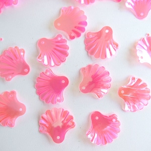 Könnte beinhalten: Rosa schimmernde Muschel-förmige Pailletten, verstreut auf einer weißen Oberfläche. Jede Paillette hat einen gewellten Rand und ein kleines Loch zum Annähen. Die Pailletten sind ein leuchtendes, durchscheinendes Rosa.