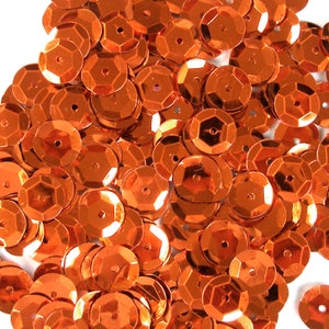 Peut inclure: Un tas de paillettes orange brillantes. Chaque paillette est un disque plat et rond avec une forme géométrique à facettes et un petit trou au centre. Les paillettes sont éparpillées, créant une surface texturée.
