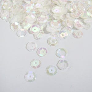 May include: A collection of iridescent white sequins, scattered across a white surface. The sequins are round with a hexagonal hole in the center, reflecting a subtle rainbow sheen. Ideal for craft projects.