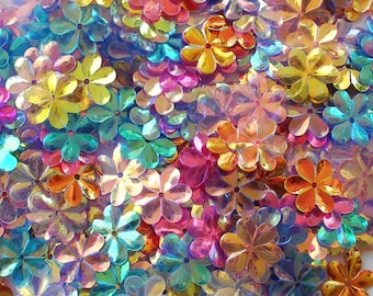 Op de afbeelding: Een verzameling iriserende bloemvormige pailletten in verschillende kleuren, waaronder geel, oranje, roze en blauw. De pailletten hebben een reflecterend, glanzend oppervlak, waardoor een glinsterend effect ontstaat. Ideaal voor knutselwerk.