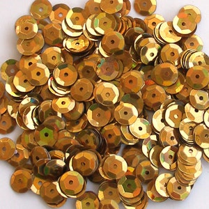 May include: A pile of shiny, golden-colored sequins. Each sequin is round with a central hole and a faceted, geometric edge. The sequins reflect light, creating a shimmering effect, ideal for embellishing crafts and clothing.