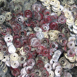 Puede incluir: Primer plano de un montón de lentejuelas decorativas metálicas. Las lentejuelas son circulares con un diseño de estrella y vienen en colores plateado y rojo oscuro. Están dispersas y superpuestas, creando una superficie texturizada.