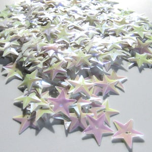 May include: A pile of iridescent star-shaped sequins. The sequins have a pearlescent sheen, with colors shifting from pale yellow to lavender and pink. Each star has five points and two small holes for sewing.
