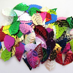 May include: A collection of colorful, metallic leaf-shaped sequins. The sequins are in various shades of green, blue, pink, red, gold, and iridescent white. Each leaf has a small hole at the top for stringing or sewing. The leaves are scattered, creating a vibrant, decorative display.