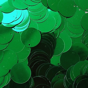May include: A close-up view of a pile of shiny, emerald green sequins. The sequins are round with a small hole near the edge, and they overlap each other. The light reflects off the sequins, creating highlights and shadows.
