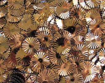 May include: A close-up shot of a pile of small, metallic gold-colored rosettes. Each rosette has a circular shape with a central hole and radiating, ridged edges, resembling miniature sunbursts. The rosettes are scattered, creating a textured, decorative effect.