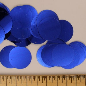 Puede incluir: Colección de lentejuelas circulares azul brillante con brillo metálico. Cada lentejuela tiene un pequeño agujero cerca del borde. Las lentejuelas están dispersas sobre una superficie neutra, con una regla visible en la parte inferior.